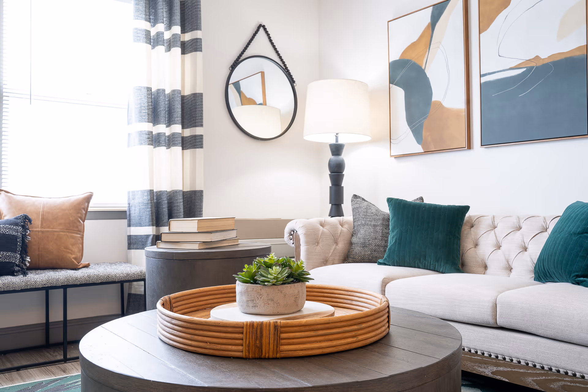 A cozy living room with a beige tufted sofa adorned with green and gray pillows, a round wooden coffee table with a decorative tray and a small potted plant, a side table with stacked books, a floor lamp, a round wall mirror, striped curtains, and two abstract paintings on the wall.