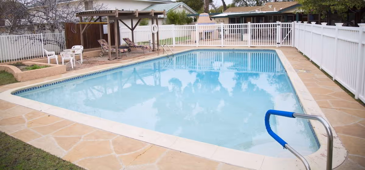 Outdoor swimming pool with clear blue water surrounded by a stone deck and white fencing. There are lounge chairs and a small shaded pergola area with additional seating on one side of the pool. Residential houses are visible in the background.