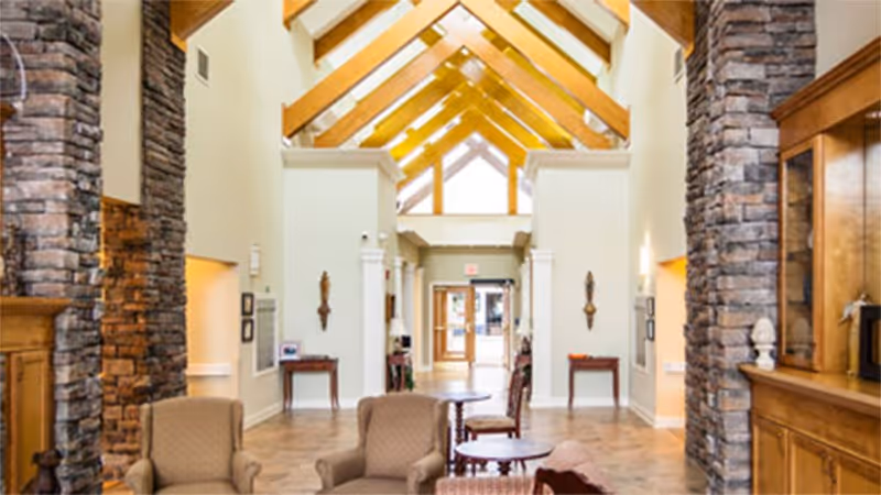 A spacious and well-lit interior common area with high vaulted ceilings featuring wooden beams and large windows. The room includes comfortable armchairs and small tables arranged for seating, stone pillars, and wooden cabinetry, creating a warm and inviting atmosphere.