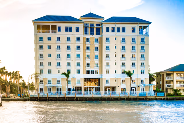 Front facade of an eight-story waterfront residential building with balconies, palm trees, and a riverside deck.