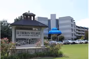 Exterior view of Lutheran Village senior living facility with a large sign in the foreground displaying the name and phone number. The multi-story building is visible in the background with several parked cars and a well-maintained lawn.