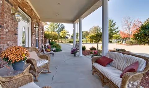 Covered outdoor patio area with wicker furniture including chairs and a sofa with cushions, potted flowers, and a view of trees and a parking lot in the background.