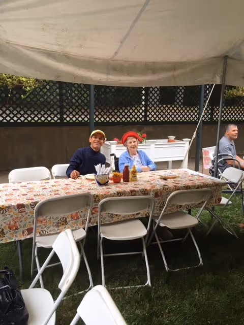 Two elderly people sitting at a long outdoor table covered with a patterned tablecloth under a canopy. The table has condiments and napkins on it. Another person is sitting in the background near a fence.