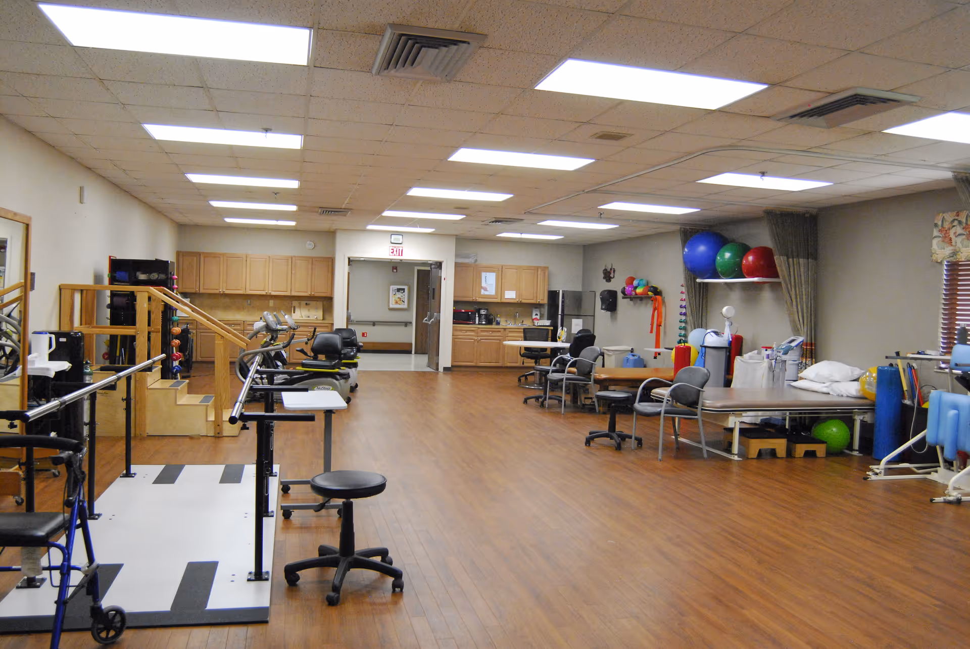 A large rehabilitation therapy room with parallel bars, exercise equipment, therapy tables, and chairs.