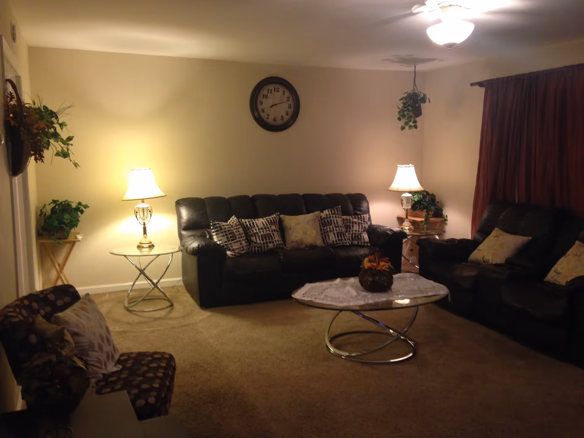 Living room with black leather sofas, armchairs, a glass-topped coffee table, table lamps, plants, and a wall clock.