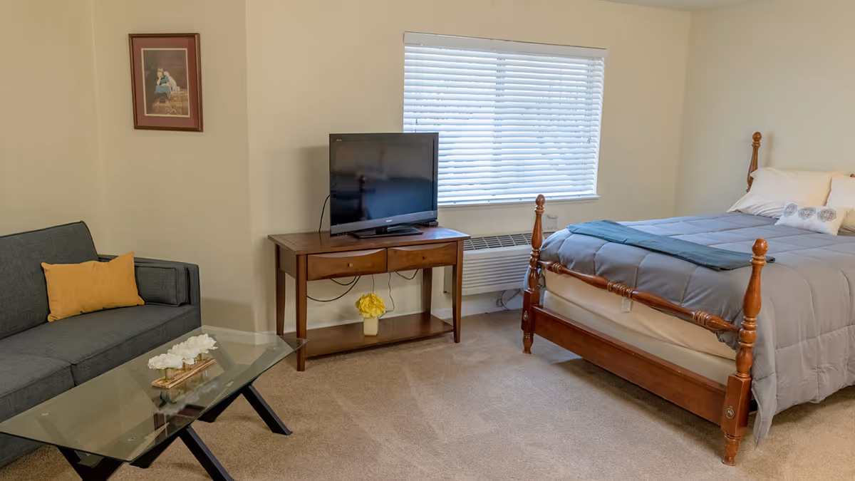A senior living community room featuring a wooden bed with gray bedding, a wooden table with a flat-screen TV, a gray sofa with a yellow pillow, a glass coffee table with white flowers, a window with blinds, and a framed picture on the wall.