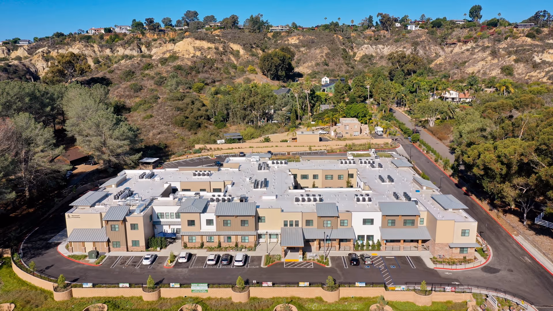 Aerial view of Westmont of Encinitas, a large senior living facility building with a white roof and beige exterior, surrounded by parking spaces and natural landscape with hills and trees in the background under a clear blue sky.