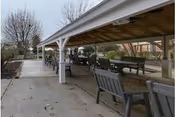 Outdoor covered seating area with benches and tables on a concrete patio, surrounded by leafless trees and a fenced garden area in the background.