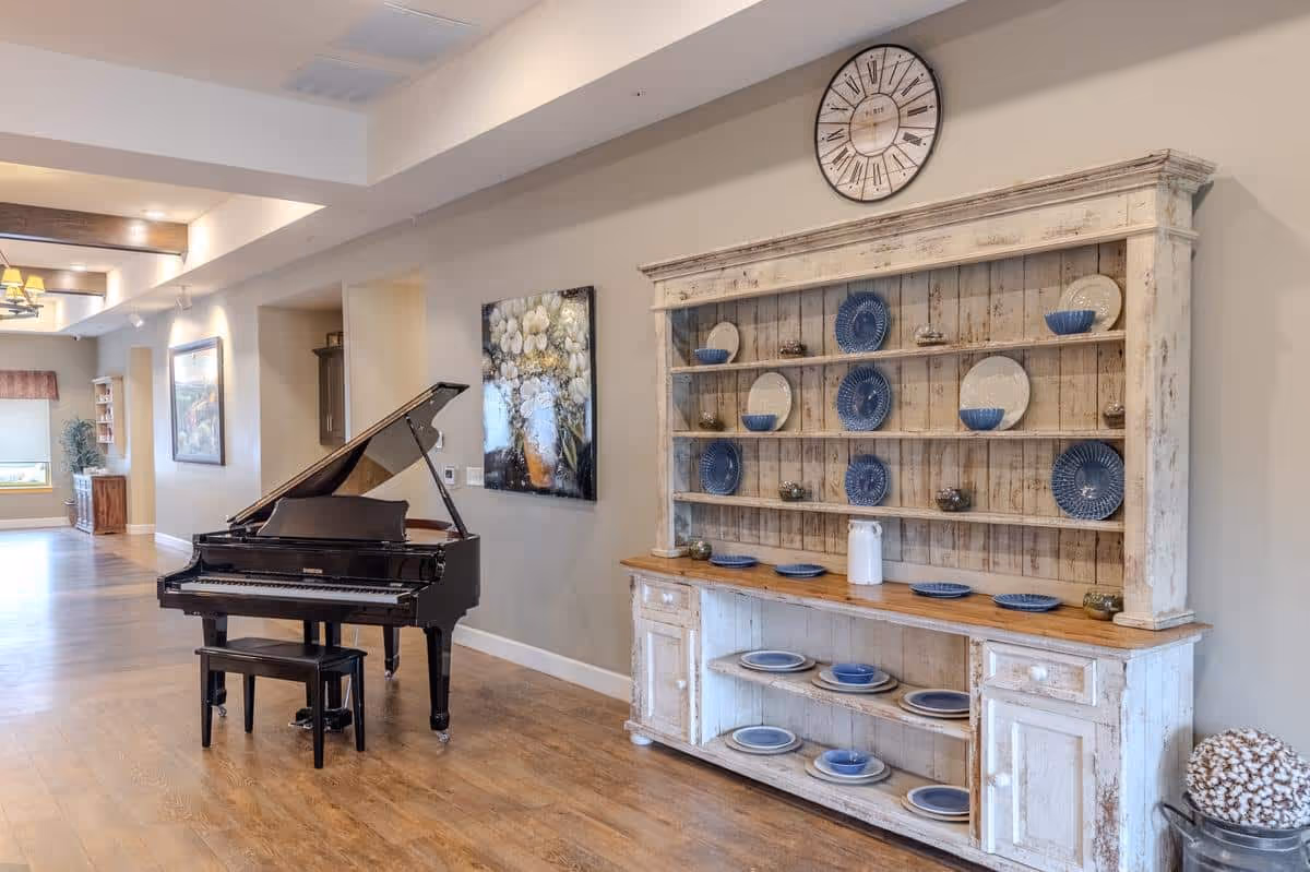 A spacious interior room with a polished wooden floor featuring a black grand piano with a matching bench on the left side. On the right side, there is a rustic white wooden hutch displaying blue and white plates and bowls. Above the hutch, a large round clock is mounted on the wall. The room is softly lit with natural light coming from windows in the background and has neutral-colored walls with some artwork hanging.