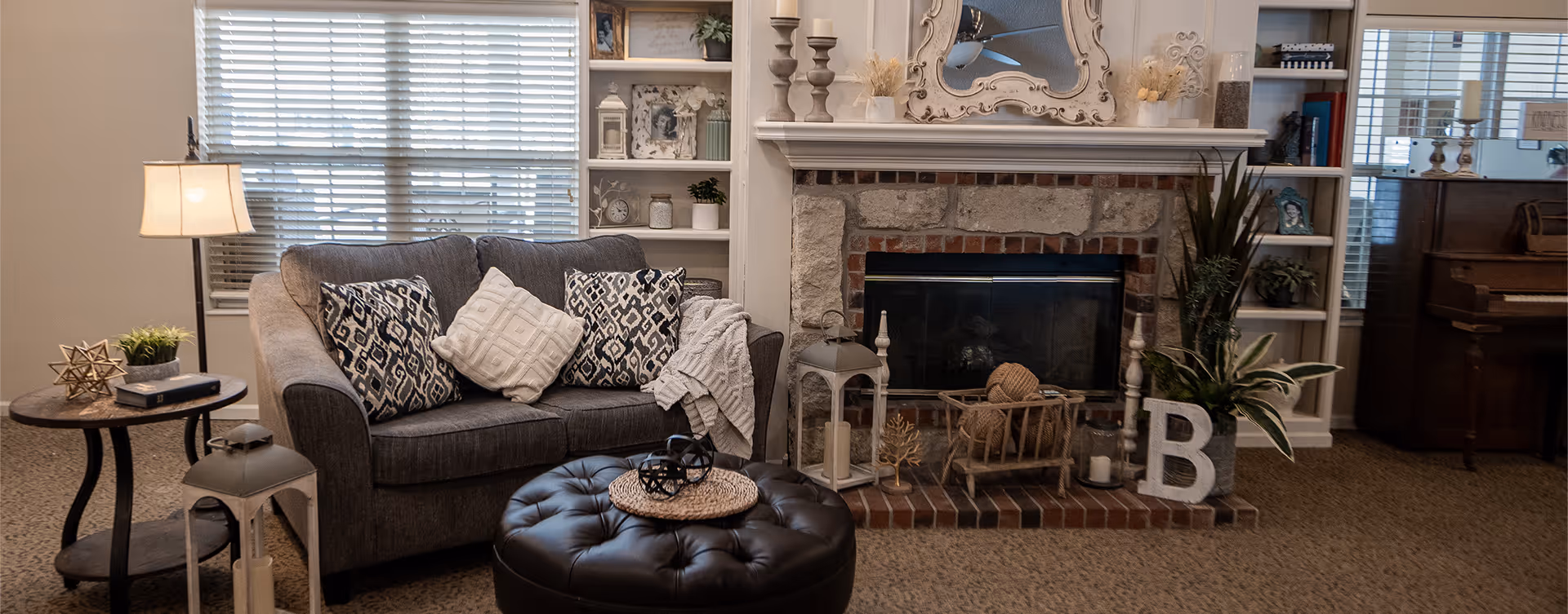 Cozy living room with a gray sofa, tufted ottoman, and a stone fireplace flanked by built-in shelves and decorative accents.