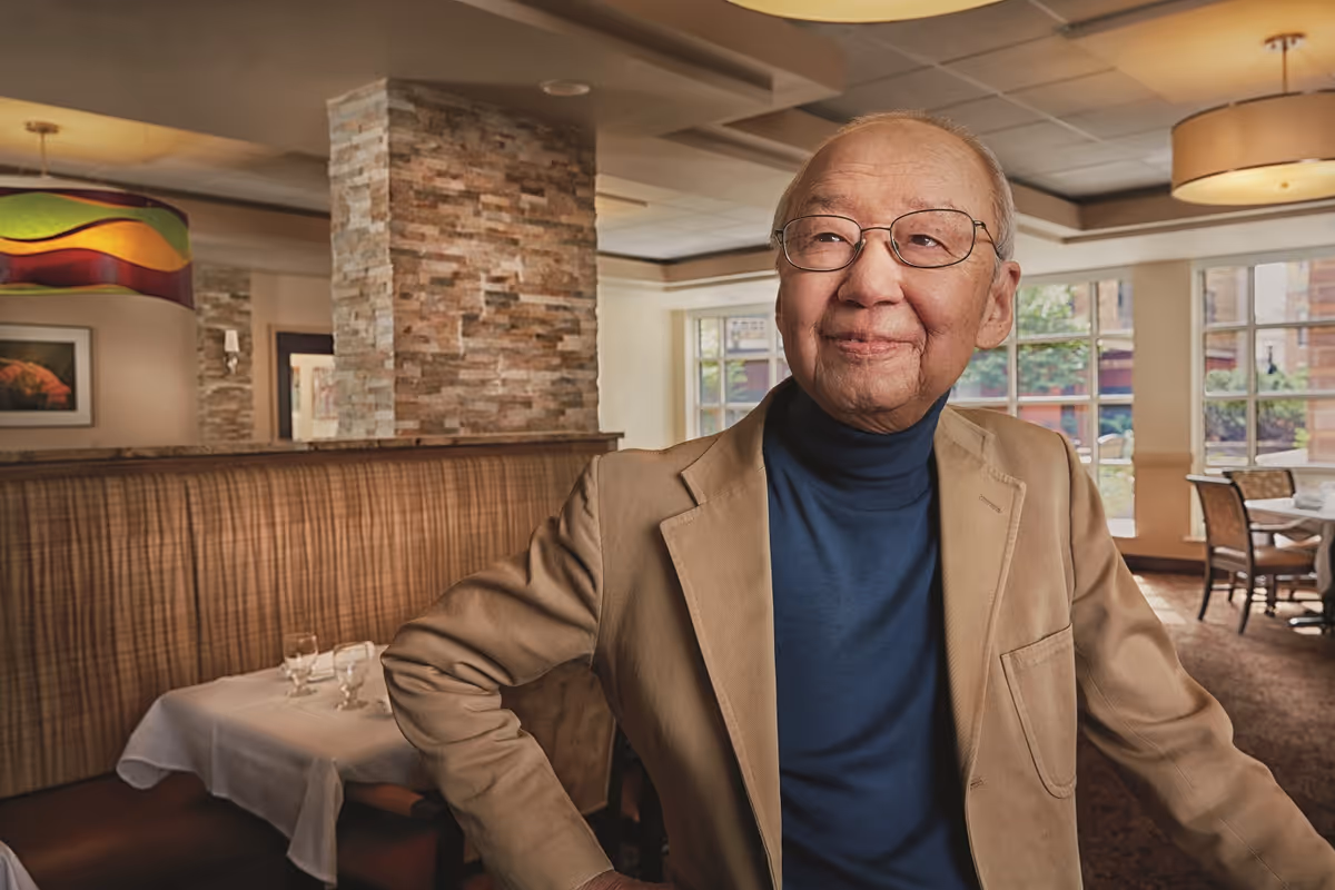 An older man wearing glasses and a tan jacket stands smiling in a well-lit dining area with tables and booth seating.