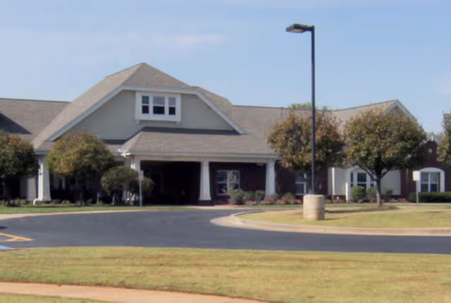 Exterior view of a single-story assisted living facility building with a sloped roof, several windows, and a covered entrance. There are trimmed trees and a paved driveway in front of the building under a clear sky.