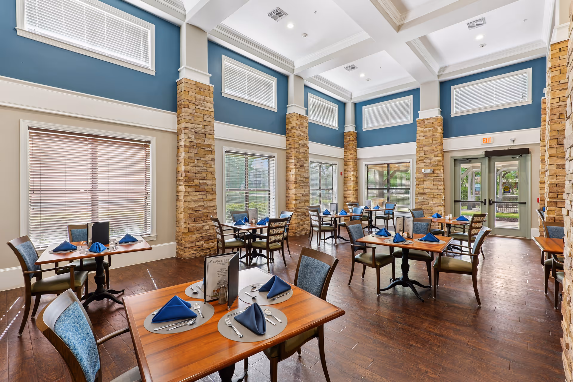 Bright dining room with set wooden tables and chairs, high coffered ceilings, stone columns, and large windows.