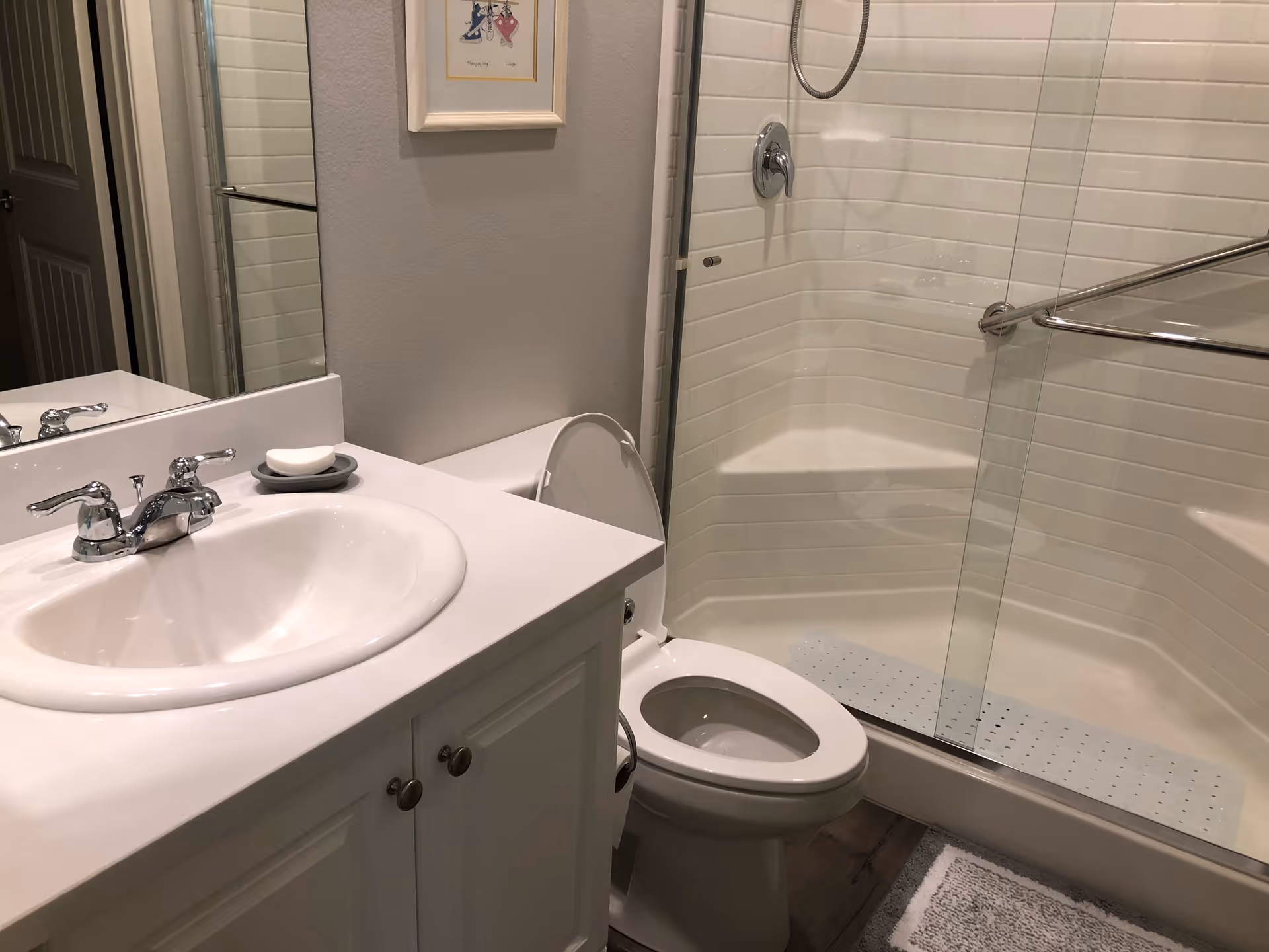 A clean bathroom featuring a white sink with chrome faucet, a bar of soap on a soap dish, a toilet with the lid open, and a glass-enclosed shower with a built-in seat and a handheld showerhead. There is a framed picture on the wall above the toilet and a gray bath mat on the floor.