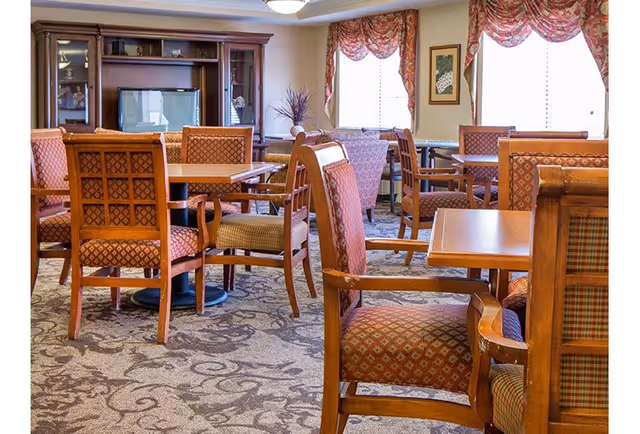 A cozy dining area with multiple wooden tables and cushioned chairs featuring patterned upholstery. The room has large windows with floral valances allowing natural light to enter. A wooden entertainment center with a television is visible against the wall, and the carpet has a decorative swirling pattern.