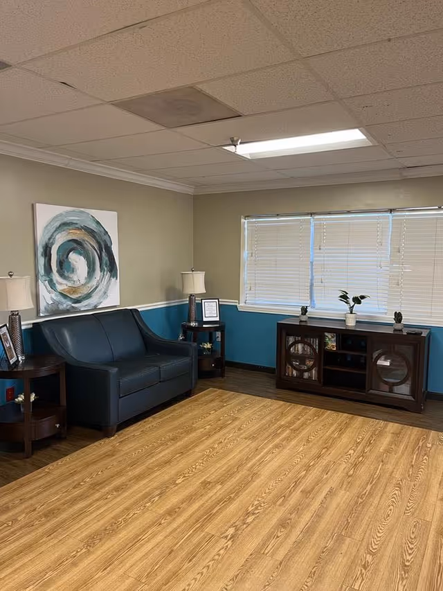A small lounge with a dark loveseat, side tables and lamps, abstract wall art, a console under a window with blinds, and wood-look flooring.