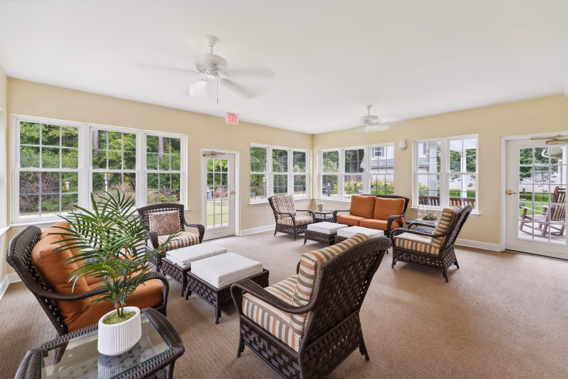 Sunlit common sitting room with wicker chairs, sofas, tables, and large windows overlooking greenery.