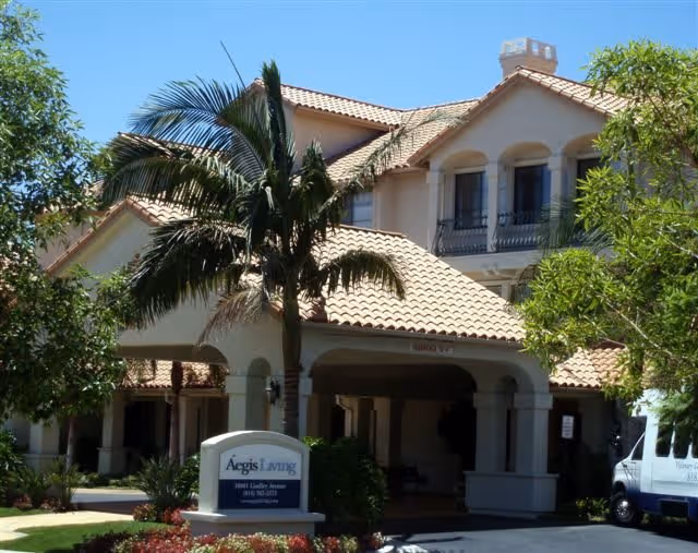 Exterior view of Aegis of Granada Hills facility showing a Mediterranean-style building with a tiled roof, palm trees, and a covered entrance. There is a sign in front with the facility name and address, and a white van is partially visible on the right side.