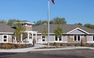 Exterior view of a single-story senior living facility building with gray siding and stone accents, a flagpole with an American flag in front, small trees, and a clear blue sky.