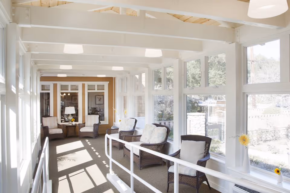 A bright sunroom with large windows on both sides letting in natural light. The room features several cushioned wicker chairs arranged along the windows, a small table with a lamp and a sunflower, and a white railing along the walkway. The ceiling has exposed white beams and modern light fixtures.