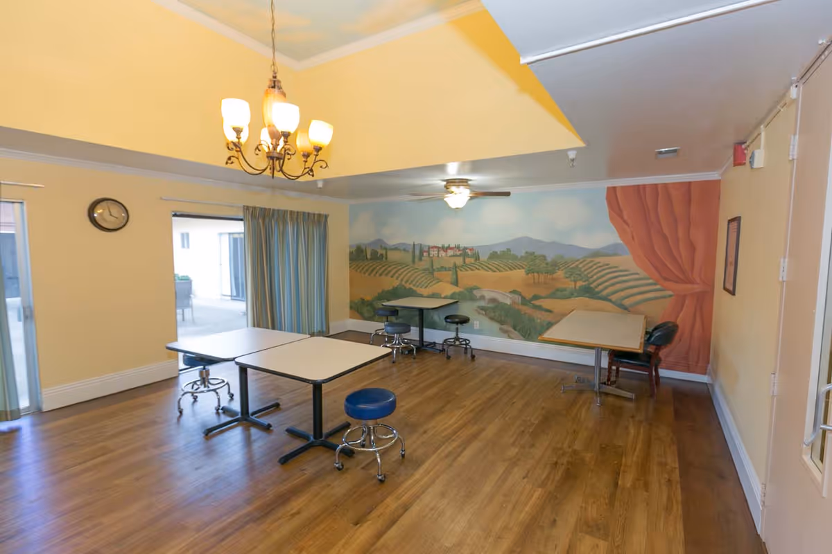 A room with wooden flooring and yellow walls featuring a mural of a countryside landscape. The room contains several tables with blue stools on wheels. There is a chandelier hanging from the ceiling and a ceiling fan with a light. A clock is mounted on the wall near a sliding glass door with blue curtains.