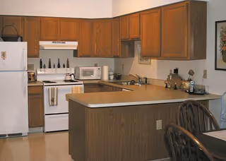 A kitchen area with wooden cabinets, a white refrigerator, a white stove with a range hood, a microwave, and a countertop with a sink. There are various kitchen items on the counters and a dining table with chairs partially visible in the foreground.