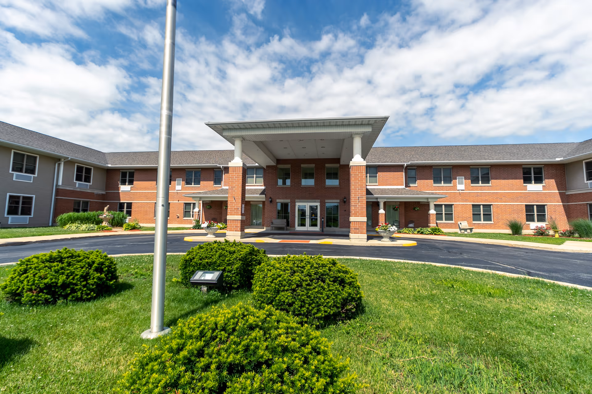 Front exterior view of a two-story senior living facility with a covered entrance supported by brick columns. The building has red brick and beige siding, multiple windows, and a circular driveway with green bushes and a flagpole in the foreground under a partly cloudy sky.