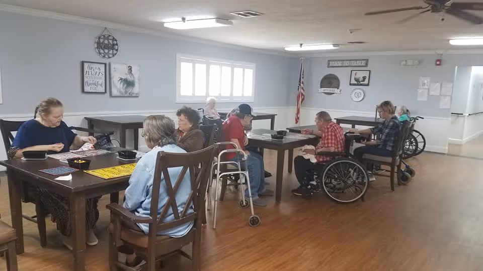 A group of elderly people sitting around tables in a well-lit room with wooden floors and light gray walls. Some individuals are in wheelchairs or using walkers. The room has several framed pictures on the walls, an American flag, and ceiling fans. The atmosphere appears to be calm and social.