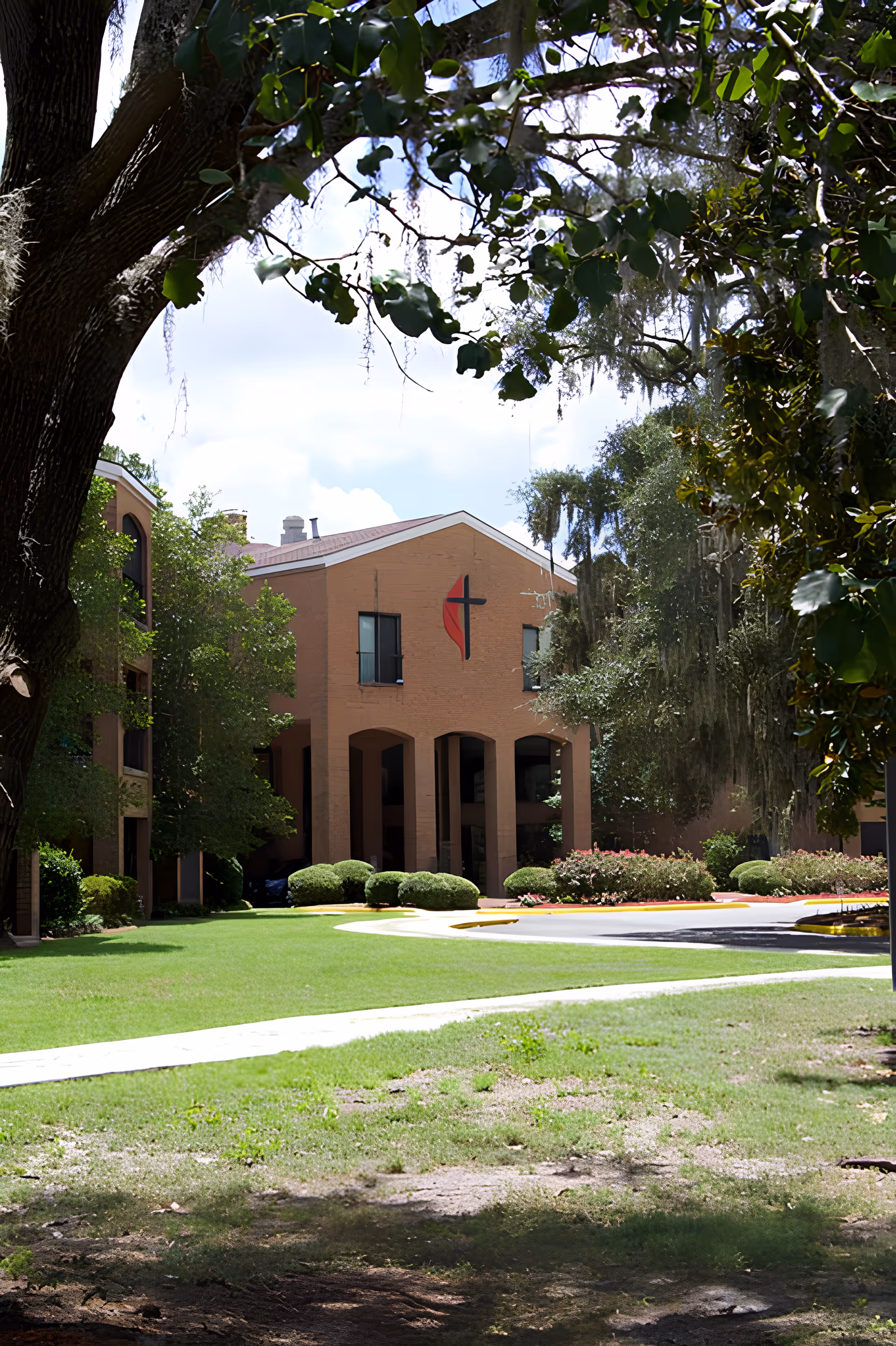 Exterior view of a brick building with a cross symbol on the front, surrounded by green trees and a well-maintained lawn under a partly cloudy sky.