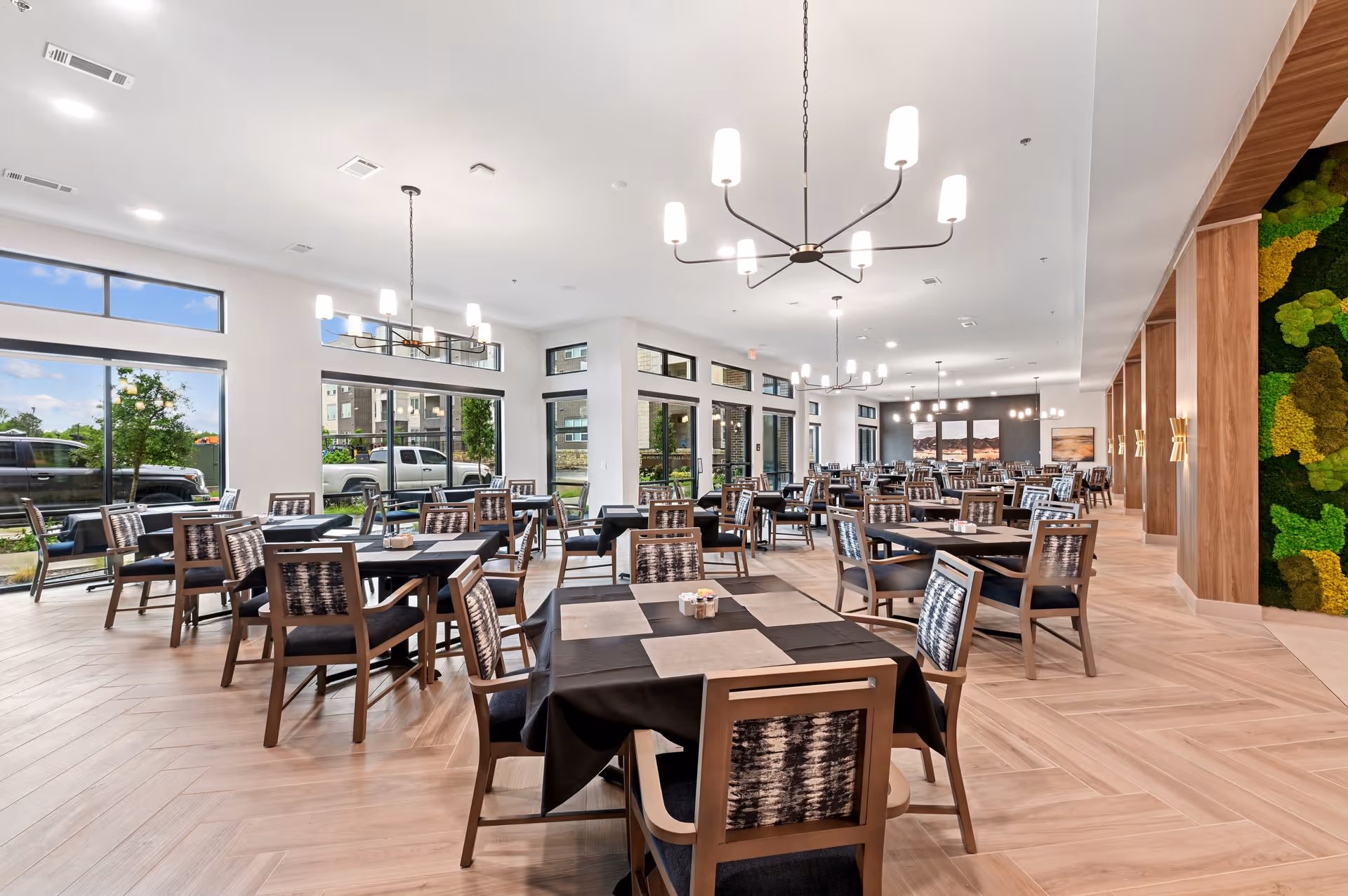 A spacious and modern dining room with multiple tables and chairs arranged neatly. Large windows allow natural light to fill the room, and contemporary chandeliers hang from the ceiling. The floor has a light wood pattern, and there is a green moss wall feature on the right side.