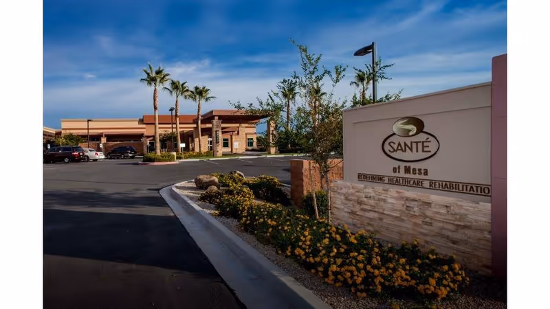Exterior view of Santé of Mesa Skilled Nursing & Rehabilitation facility with a clear blue sky, palm trees, landscaped flower beds, and a parking area with several cars.