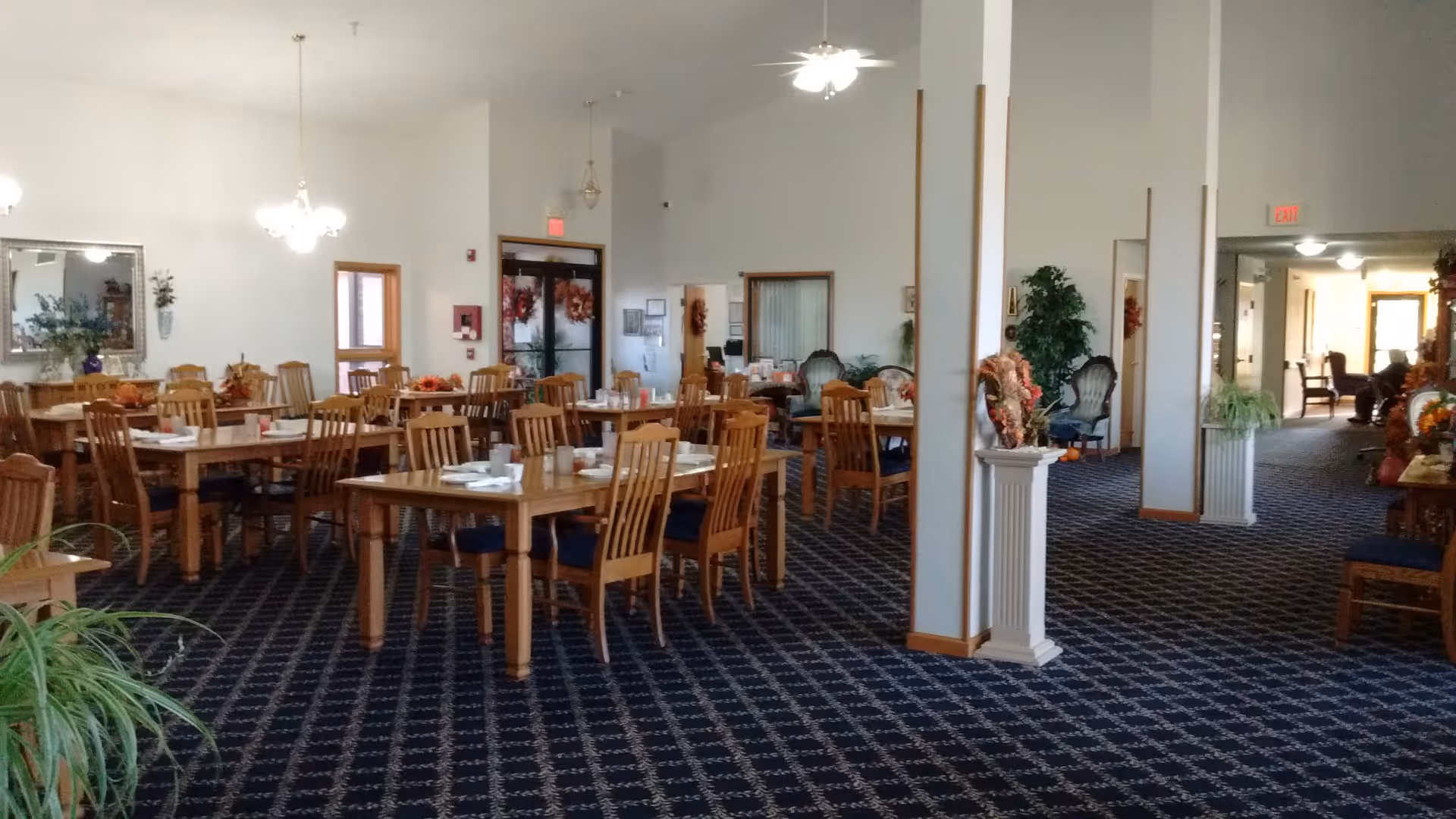 Spacious dining room with multiple wooden tables and chairs arranged neatly on a dark patterned carpet. The tables are set with white napkins and cups. The room has white walls, several ceiling lights, and decorative plants and mirrors along the walls. There is a hallway visible to the right leading to other areas.