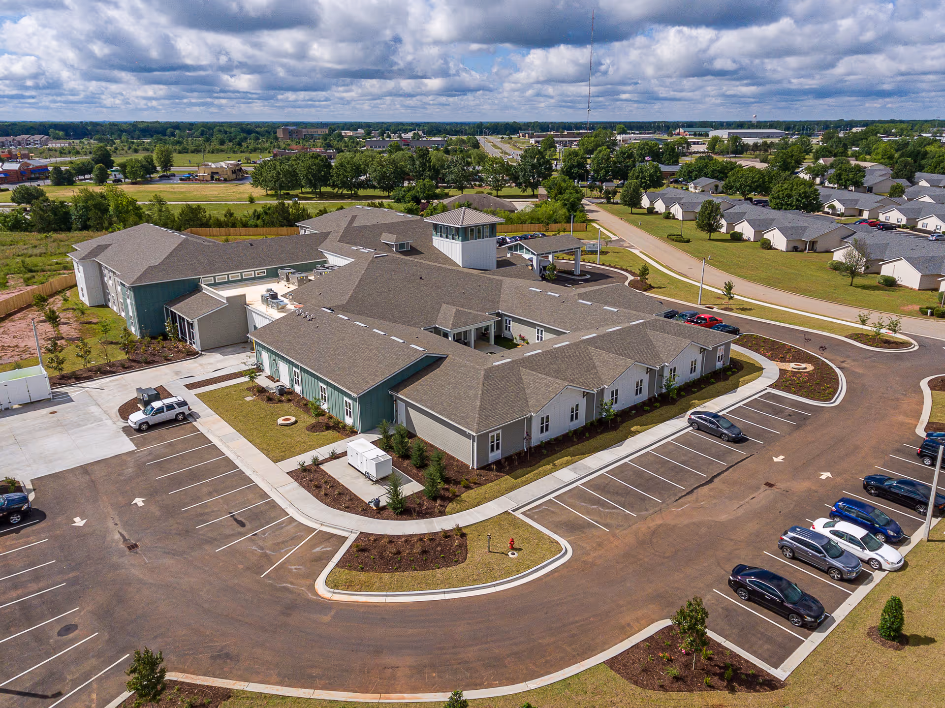 Aerial view of The Canopy at Warner Robins senior living facility showing a large single-story building with a gray roof surrounded by a parking lot with several cars. The facility is located in a suburban area with green lawns, trees, and nearby residential houses under a partly cloudy sky.