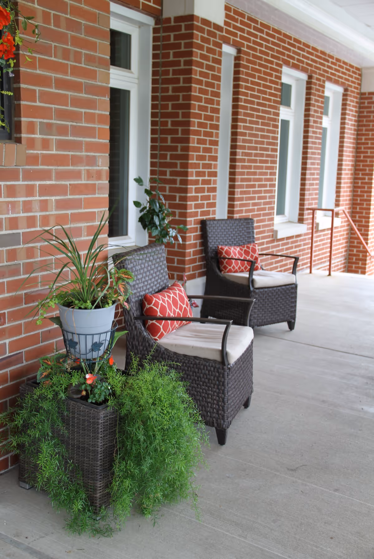 Outdoor covered patio area with two dark wicker chairs featuring white cushions and red patterned pillows, positioned against a red brick wall with white-framed windows. There are green plants in pots placed near the chairs, adding a touch of nature to the space.