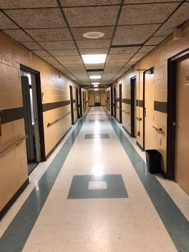 A long, narrow hallway in a facility with beige walls and a brown horizontal stripe running along the middle. The floor has a pattern of green and white tiles with square designs. Several doors line both sides of the hallway, some open and some closed. Handrails are mounted on the walls, and ceiling lights illuminate the corridor.