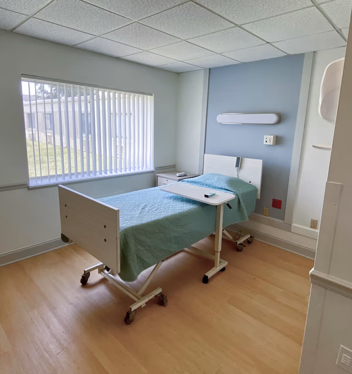 A simple patient bedroom with a single hospital bed, overbed table, bedside cabinet, and a large window with vertical blinds.