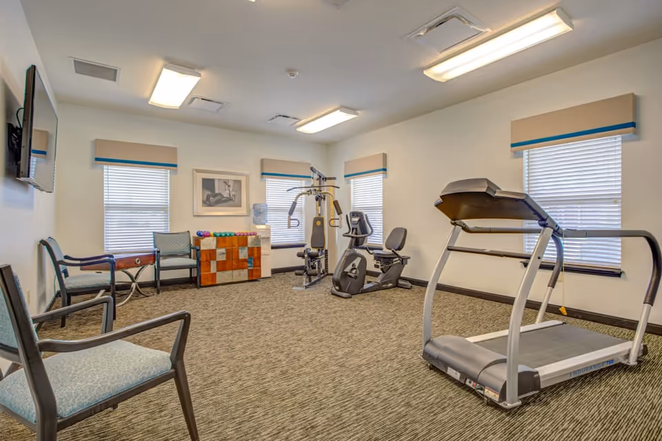 A well-lit exercise room with a treadmill, stationary bike, and multi-function weight machine. The room has three windows with beige and blue valances, a water cooler, a rack of colorful dumbbells, and several chairs along the walls. A flat-screen TV is mounted on one wall.