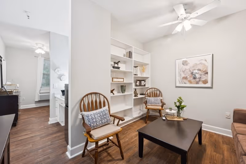 A cozy living room area with two wooden chairs featuring patterned cushions, a dark wooden coffee table with a small plant and remote control on it, a white built-in bookshelf with decorative items, a ceiling fan with lights, and a framed floral artwork on the wall. The room has wooden flooring and light-colored walls.