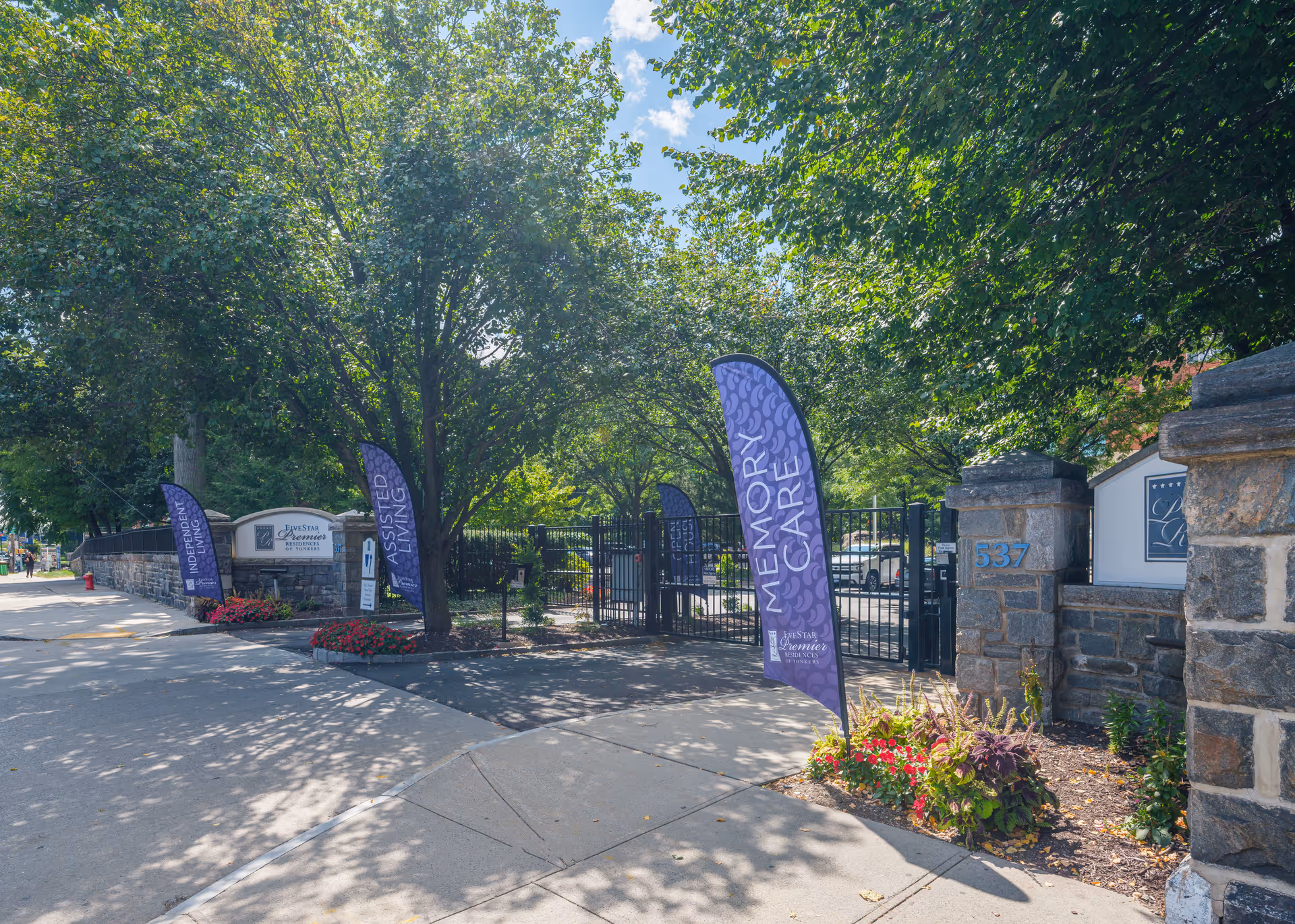 Stone entrance and gated front of a senior living facility with purple 'Memory Care' flags, signage, and trees along the sidewalk.