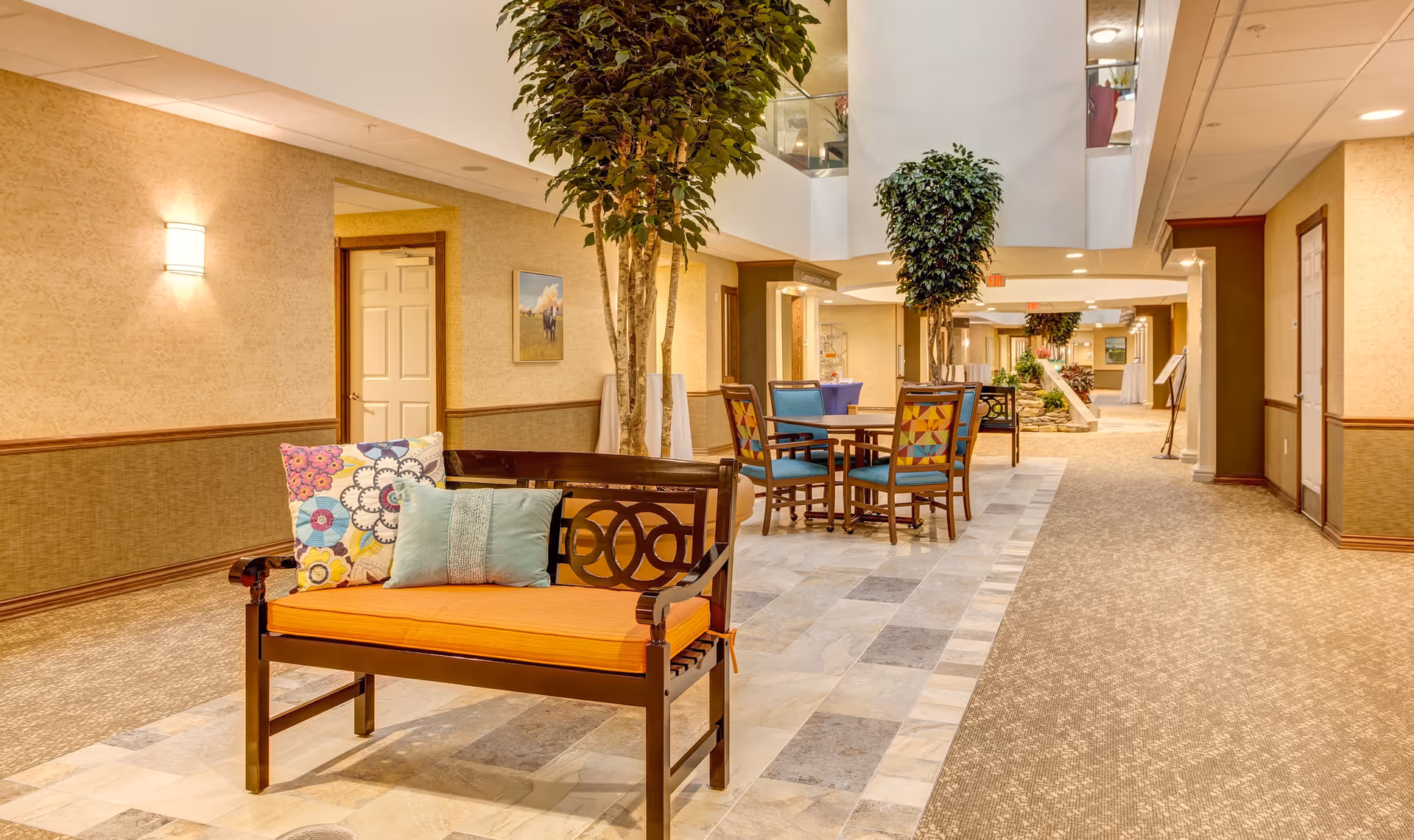 Interior view of a retirement community hallway with a wooden bench featuring orange and patterned cushions in the foreground. There are potted trees, a table with four colorful chairs, and warm lighting along the beige walls. The hallway extends into the distance with additional seating and decor visible.