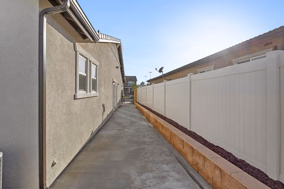 A narrow outdoor walkway between two buildings with a concrete path, beige stucco walls on the left, and a white fence on the right. There is a small garden bed with red mulch along the base of the fence. The sky is clear and sunny.