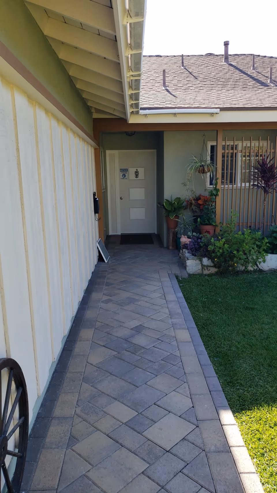 A paved walkway leading to a white door at the end of a building exterior. The walkway is bordered by a green lawn and a garden area with various potted plants and hanging plants. The building has a beige wall with vertical paneling and a sloped roof with shingles.