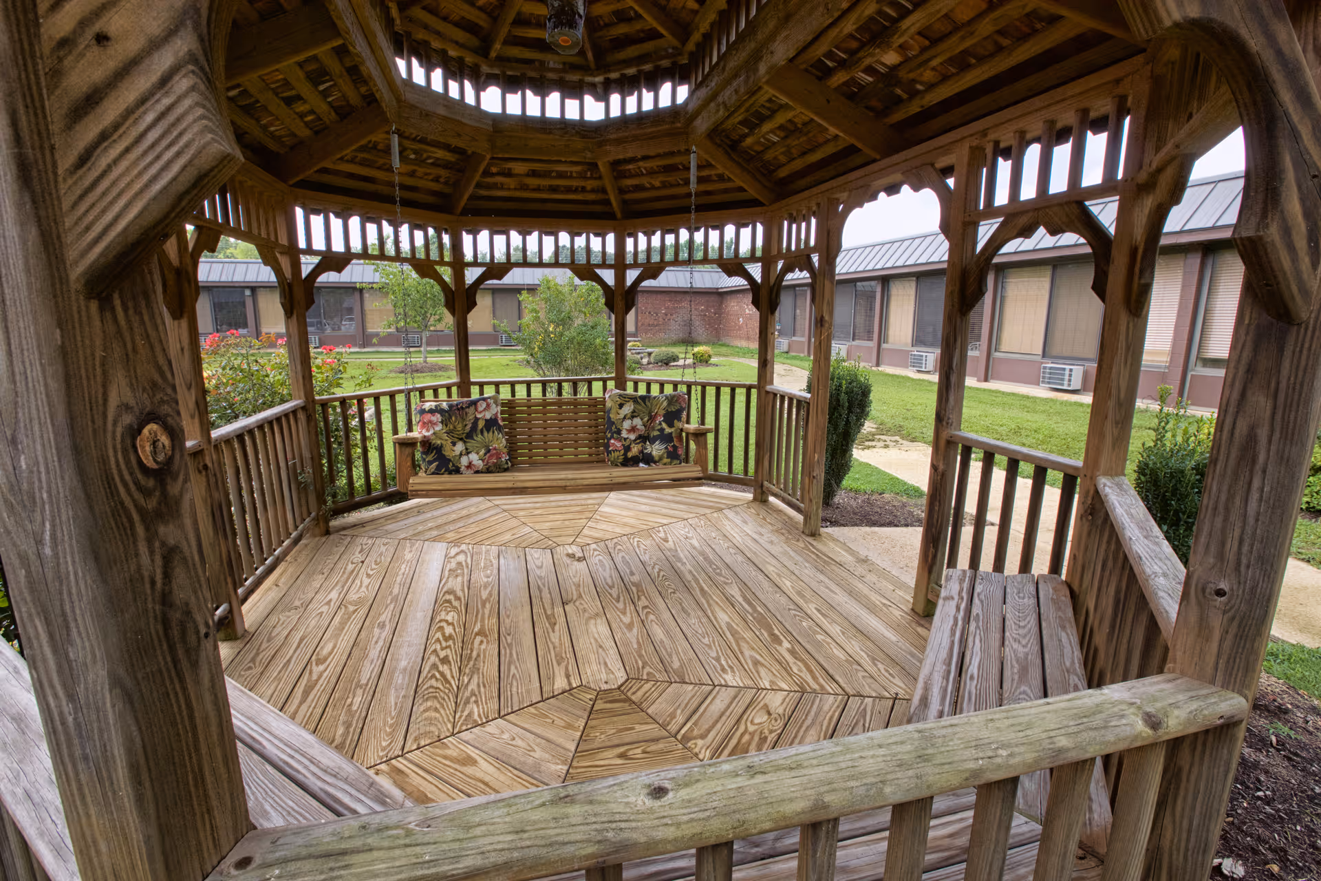 Wooden gazebo with built-in benches and a swinging bench with floral cushions overlooking a grassy courtyard and adjacent single-story building.