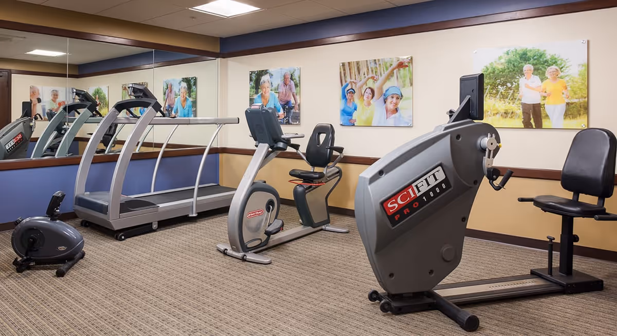 Exercise room with various fitness equipment including a treadmill, stationary bike, and rowing machine. The room has carpeted flooring, beige and blue walls, and large mirrors on one side. Several framed photos of elderly people engaging in outdoor activities are hung on the walls.