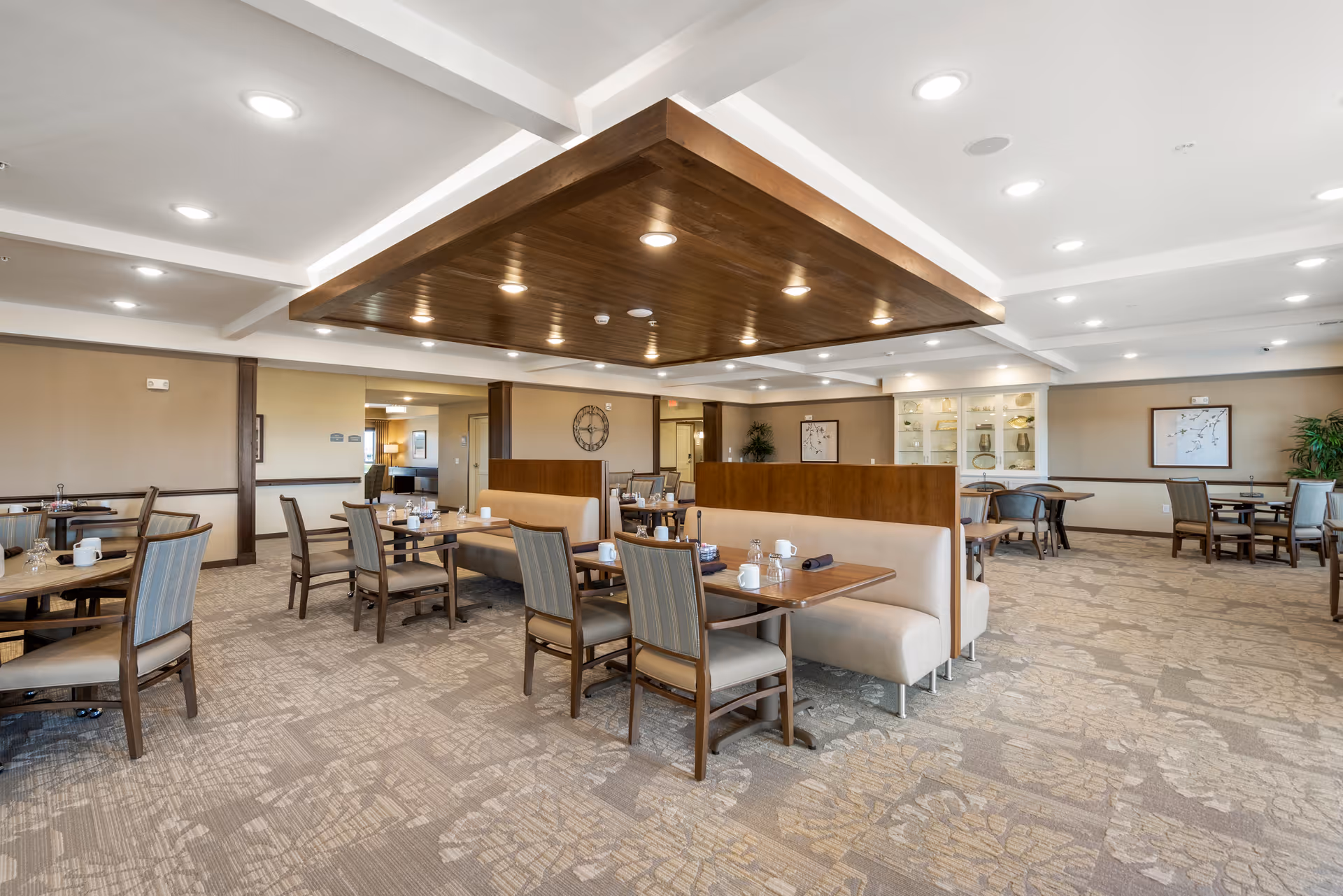 A spacious dining room in Urbana Place Senior Living featuring multiple wooden tables and chairs arranged neatly. The room has a beige and brown color scheme with patterned carpet flooring, a wooden ceiling accent with recessed lighting, and decorative wall art. The tables are set with cups, glasses, and napkins, creating a welcoming atmosphere.