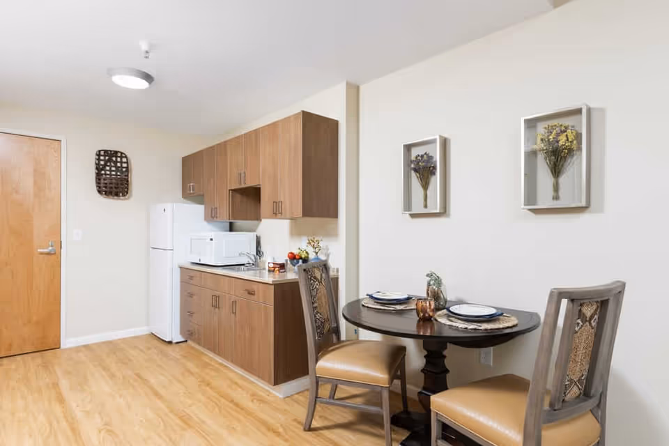 A small kitchen and dining area in a senior living facility. The kitchen features wooden cabinets, a white refrigerator, a microwave, and a countertop with a sink. Adjacent to the kitchen is a round dining table set for two with plates and placemats. Two chairs with cushioned seats are positioned around the table. The walls are decorated with two framed floral arrangements, and the floor has light wood-style flooring.