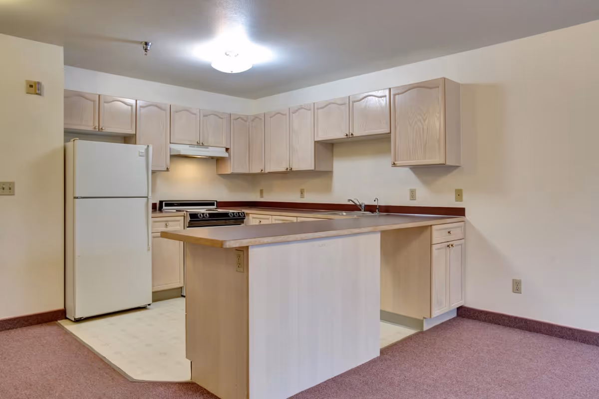 A kitchen area in a senior living facility featuring light wood cabinets, a white refrigerator, a stove with an overhead vent, a double sink, and a countertop island. The floor in the kitchen area is covered with light-colored linoleum, while the adjacent area has a mauve carpet. The walls are painted white, and a ceiling light fixture illuminates the space.