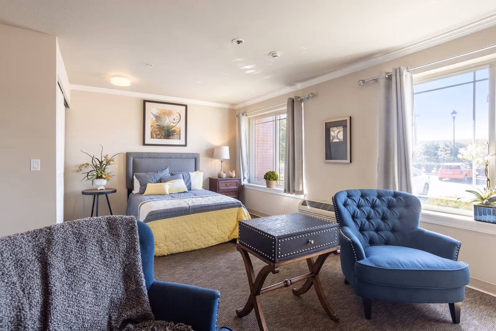 A bright and cozy bedroom in an assisted living facility featuring a bed with gray and yellow bedding, a nightstand with a lamp, a small round table with a plant, two blue armchairs, a small wooden table, and large windows with gray curtains letting in natural light.