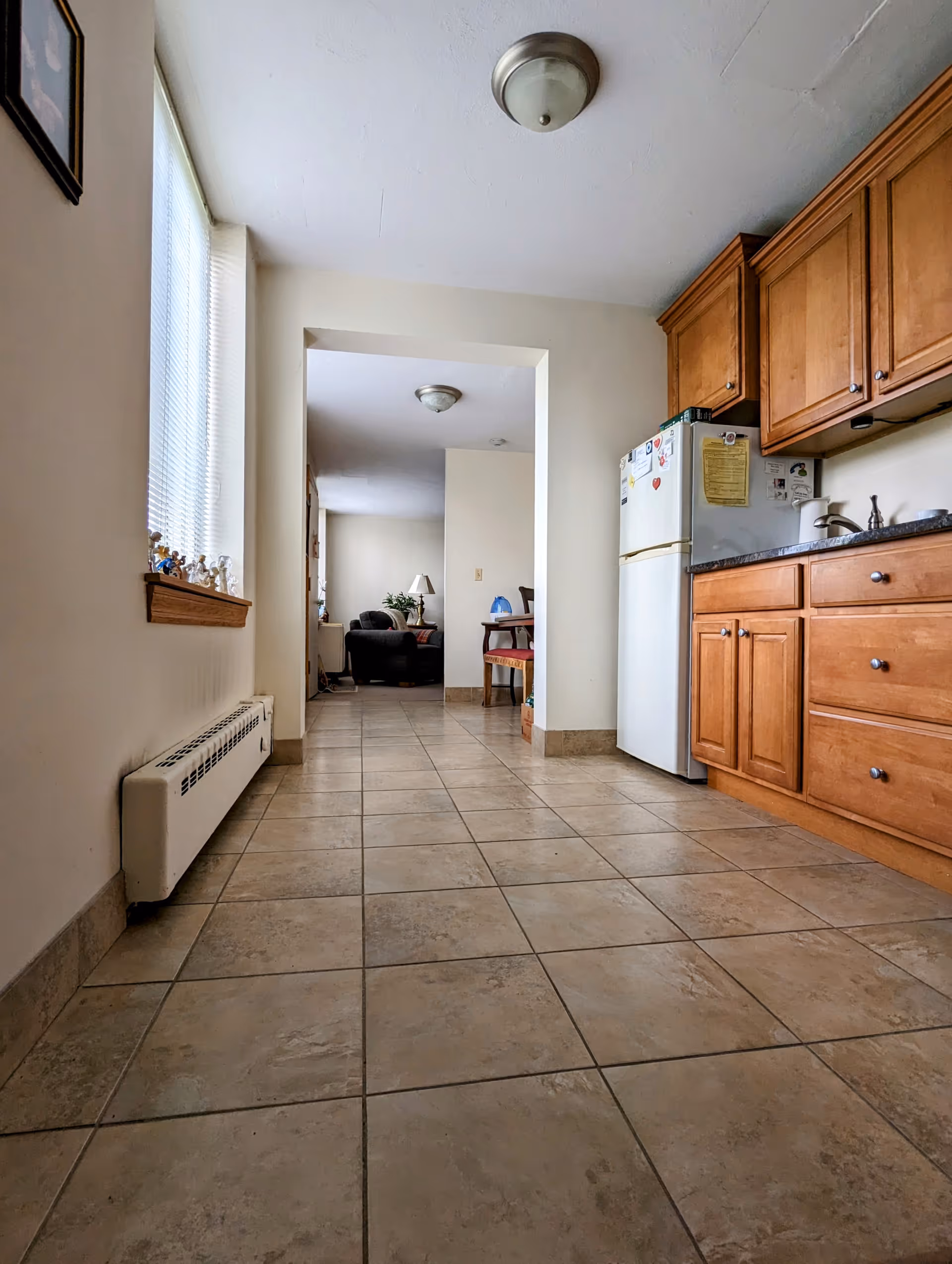 View down a tiled floor hallway leading from a kitchen area with wooden cabinets and a white refrigerator on the right to a living room area with a black armchair and a lamp in the background. There is a window with blinds on the left wall and a heating unit below it.