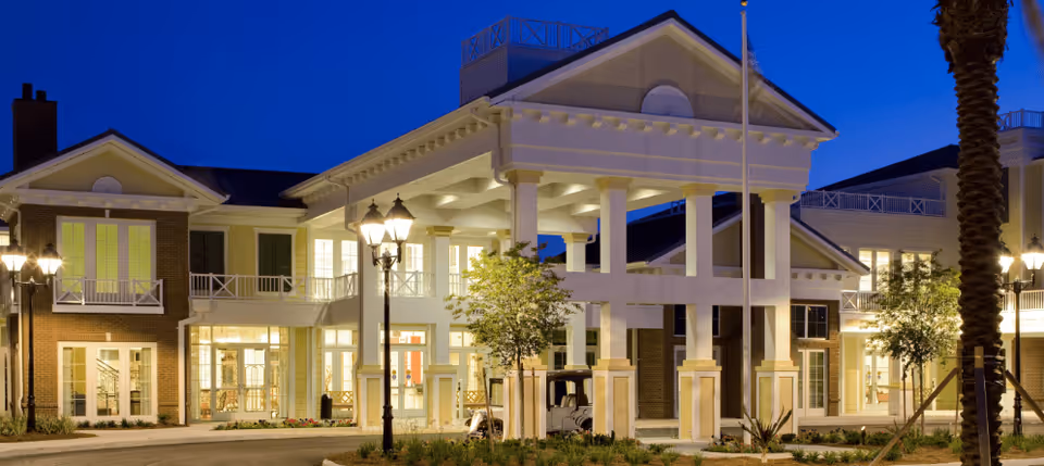 Front entrance of a two-story residential building with a large columned portico and illuminated windows at dusk.